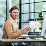 Smiling young boss sits at a desk in front of a computer, makes a video call presentation online, shows graphs, looks at the camera and smiles.